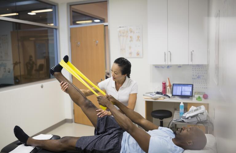 Male African American patient using stretch band on leg in physical therapy with female Asian American therapist