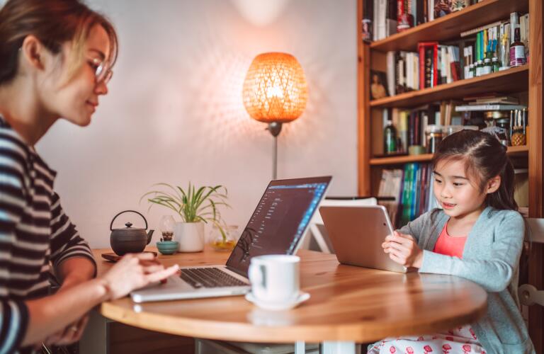 Asian mother working on laptop at desk with young daughter looking at tablet