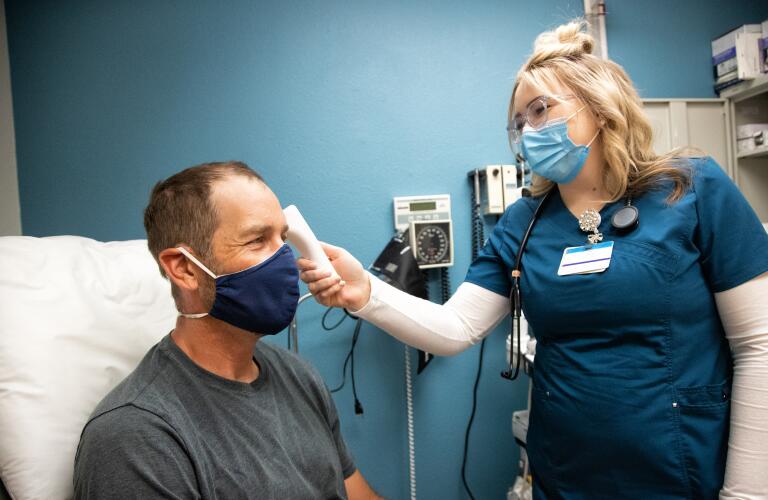 young female nurse wearing surgical mask takes temperature of masked white male in his forties in an examination room of a medical clinic