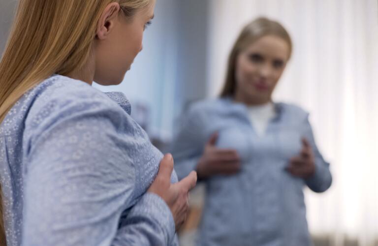 Young female touching chest in front of mirror, unsatisfied with her breast size