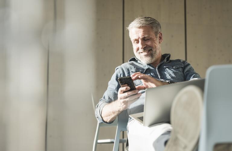 middle age man smiling at smartphone with laptop, working or socializing