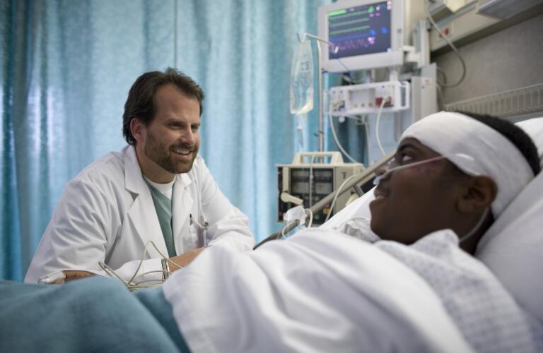 African American male patient with head bandage in hospital bed talking to Caucasian male doctor