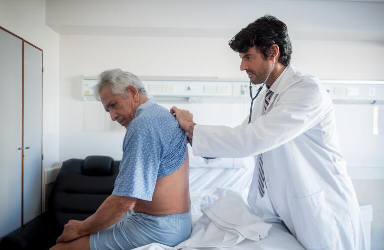 Caucasian male doctor checking breathing of older Caucasian patient with stethoscope on patient's back