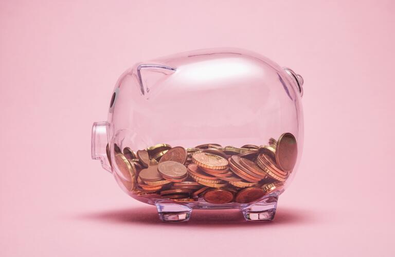 Clear piggy bank filled with coins against pink background