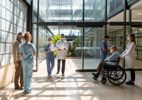 hospital staff with surgical masks walking in and out of the hospital, with a male patient in wheelchair being wheeled into the hospital