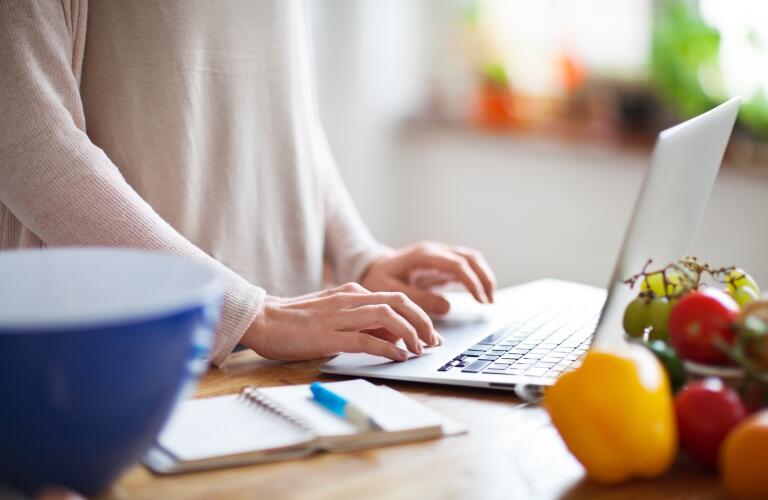 Woman looking something up online using a laptop computer in kitchen, with notebook and pen nearby