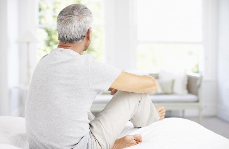 back view of senior man sitting on bed and looking out window