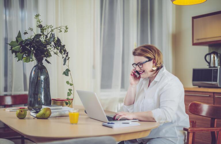 Young Caucasian overweight woman on phone in kitchen looking at laptop smiling