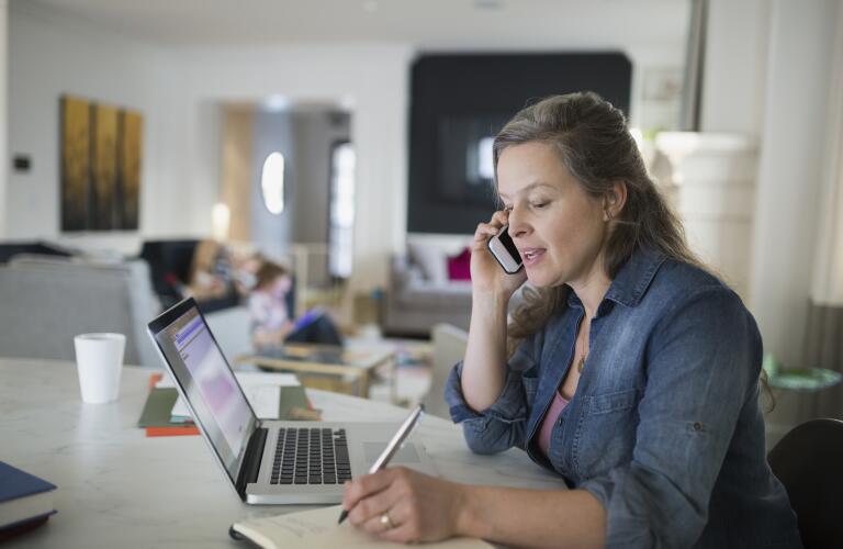 middle aged Caucasian woman taking notes on paper and talking on smartphone in front of laptop