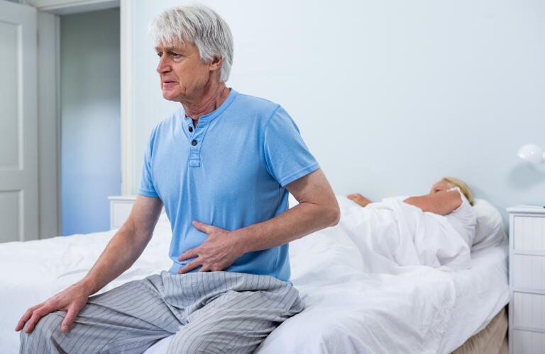 senior man sitting up in bed with hand on stomach