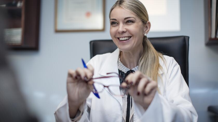 portrait of smiling Caucasian female eye doctor at desk holding eye glasses and pen