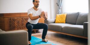Muscular Athletic Fit Man in T-shirt and Shorts is Doing Squat Exercises at Home