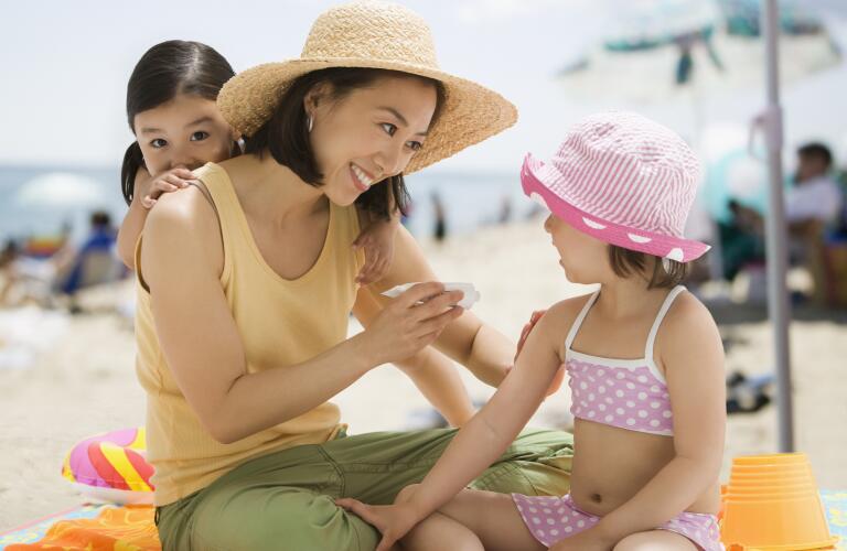 Asian mother wearing sun hat applying sunscreen on children at beach