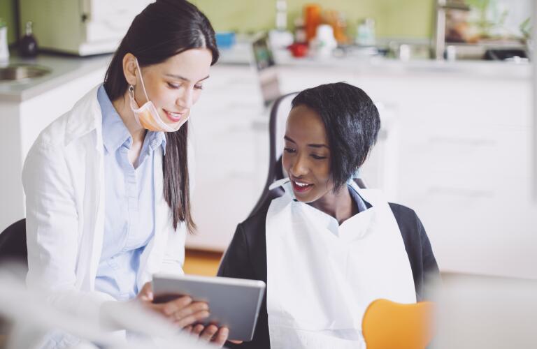 Young female African American patient looking at tablet with young female Caucasian dentist or hygienist