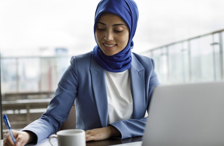 young woman in head scarf at a cafe with laptop, pen and paper, and cup of tea or coffee