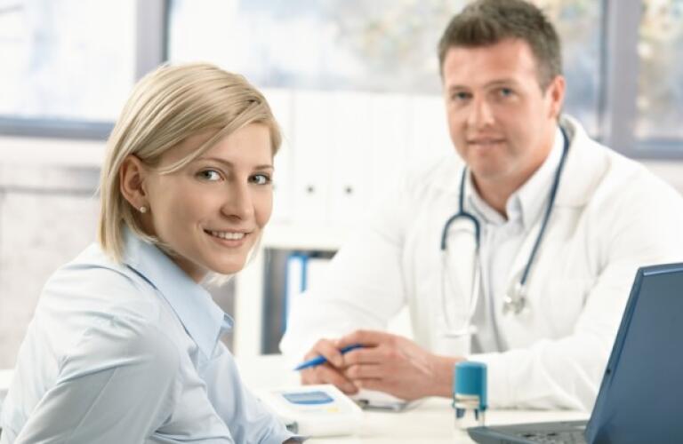 Smiling women sitting in doctors office on appointment looking at camera with doctor in background