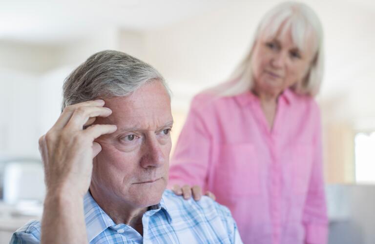 Senior Caucasian male looking concerned or stressed while senior Caucasian woman comforts him in background