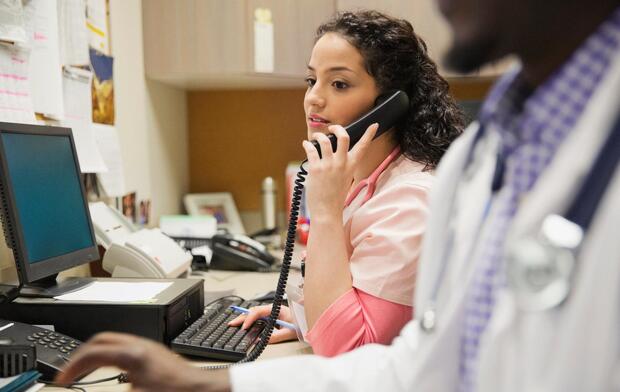 Female medical professional using telephone while working at desk with colleague in foreground